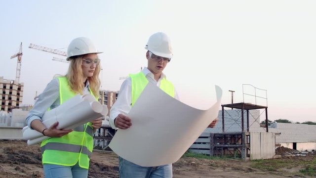 Workers with drawings at the construction site. Two workers man and woman in protective harhats working with drawings at the construction site outdoors
