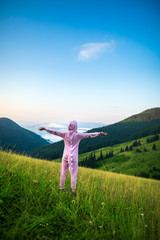 Positive young woman hiker in pink Dragon Pajamas open arms at mountain peak. Slim lady in pink night-suit. Funny travel, Animal Cosplay Costume. Summer adventure journey in mountain nature outdoors