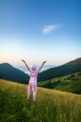Positive young woman hiker in pink Dragon Pajamas open arms at mountain peak. Slim lady in pink night-suit. Funny travel, Animal Cosplay Costume. Summer adventure journey in mountain nature outdoors - 284524350