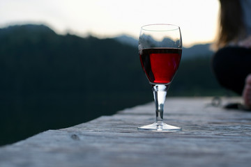 Ein Glas Rotwein auf einem Holzsteg am See mit verschwommener Person im Hintergrund