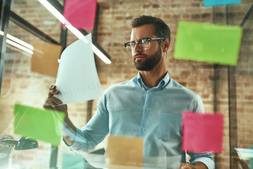 Creativity. Young and handsome bearded man in eyeglasses planning working process and using colorful stickers while standing in front of glass wall