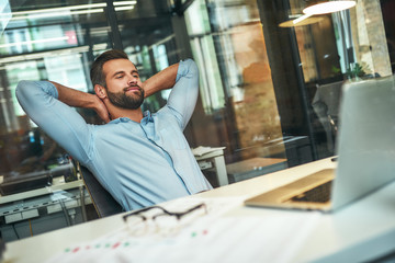 Work done Satisfied young bearded businessman leaning back with hands behind head and smiling while sitting in the modern office