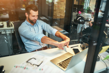 Relaxing at workplace. Tired young bearded businessman in formal wear stretching his arms and smiling while sitting in the modern office