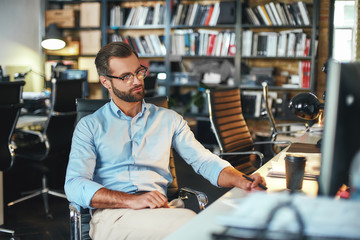Concentration. Young bearded businessman in eyeglasses and formal wear working on computer and thinking about something while sitting in modern office