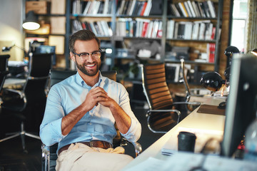 Smart and successful. Young and cheerful bearded man in eyeglasses and formal wear working on computer and smiling while sitting in modern office
