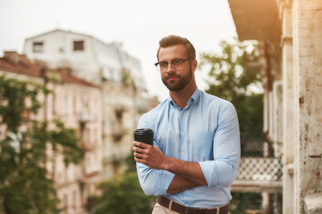 Relaxing after work. Young and handsome bearded man in eyeglasses and formal wear holding cup of coffee and looking away while standing at the office balcony