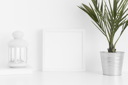 White square frame mockup with a palm in a pot, candle holder and a book on a white table.