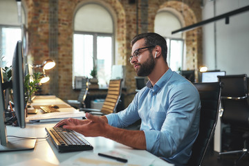 I don't understand. Side view of young and successful bearded man in eyeglasses and headphones typing something on computer and gesturing while sitting in the modern office