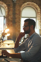 Working in the evening. Side view of young and successful bearded man in eyeglasses and formal wear looking at computer monitor while sitting in the modern office