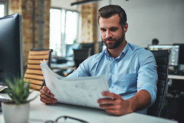 Great work Concentrated bearded engineer in formal wear looking at blueprint and smiling while sitting in the modern office