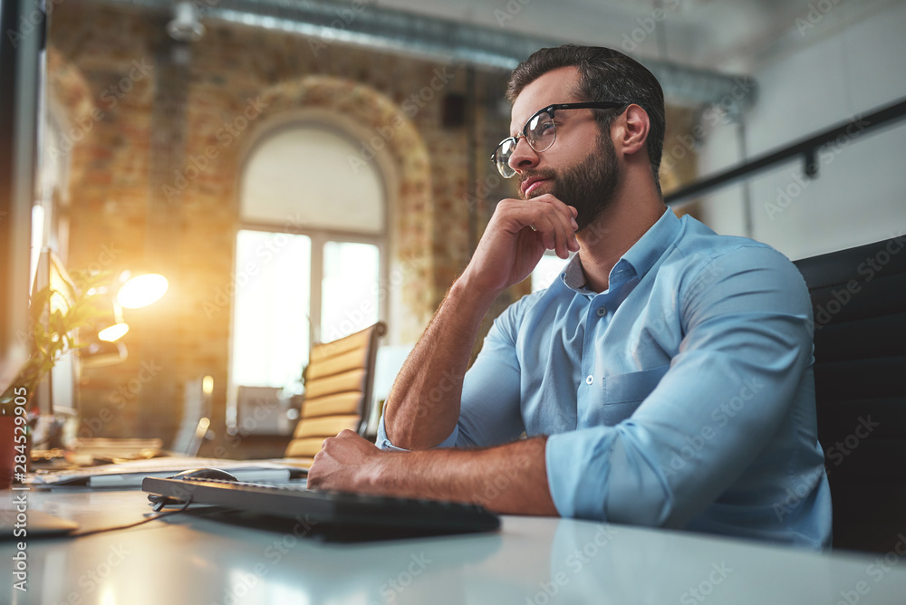 Wall mural Thinking about... Side view of young bearded man in eyeglasses and formal wear working on computer and touching his chin while sitting in the modern office