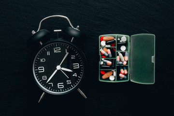 Old alarm clock wut green pill case on black desk as backgrounds - vintage bell clock and medicine organizer placed on dark stone surface