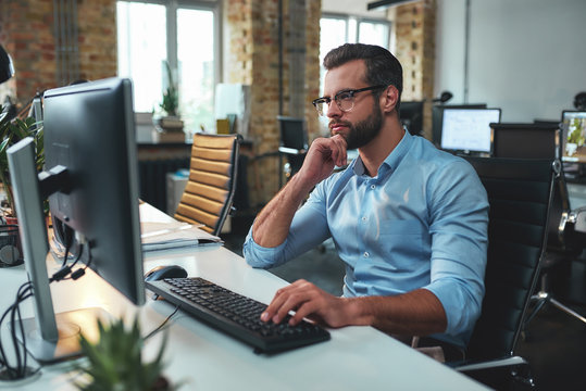 New project. Side view of young bearded man in eyeglasses and formal wear working on computer and touching his chin while sitting in the modern office