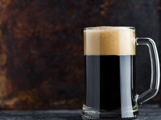 Glass mug of dark beer with foam cap on rustic rusty background