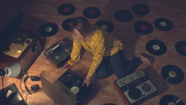 Girl surrounded by vintage devices for reproducing and recording sound.  Girl sits on the floor. Next to her are bobbin tape recorders of the 60s, 70s and 80s, an old turntable and records.