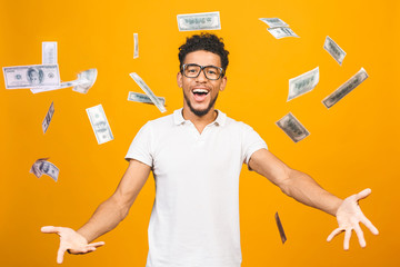 Portrait of a happy young afro american man throwing out money banknotes isolated over yellow...