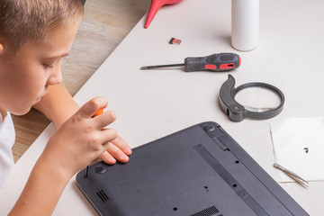 A boy of 10 years old is sorting a laptop for cleaning and maintenance. Selective focus. Screwdrivers, purge cylinder, magnifying glass and spray cleaner in the frame.