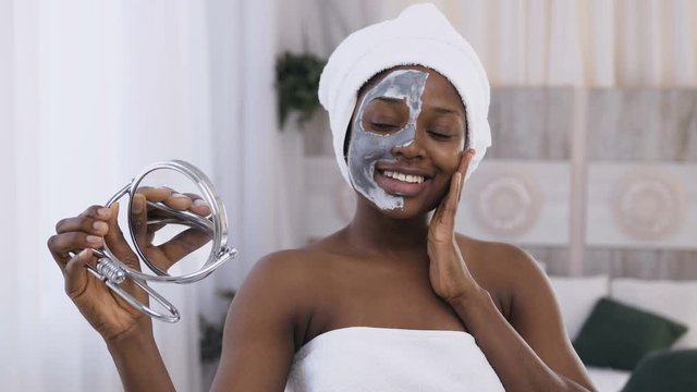 Portrait of young smiling african woman with white towel on head applied clay mask on half face and watching in the mirror