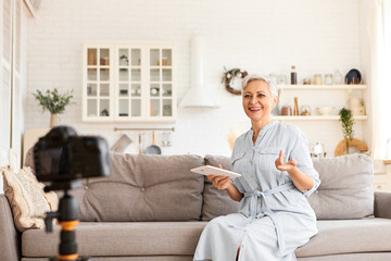 People, technology and modern electronic gadgets concept. Picture of beautiful short haired senior female blogger sitting on couch in kitchen interior, using digital tablet in front of camera tripod