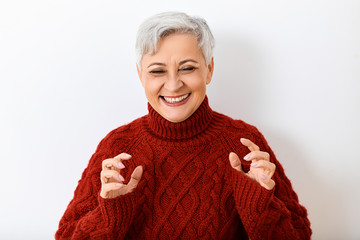 Joyful happy elderly sixty year old female wearing stylish maroon turtleneck pullover laughing out loud, having fun, posing isolated holding hands like claws. Sincere emotions and body language