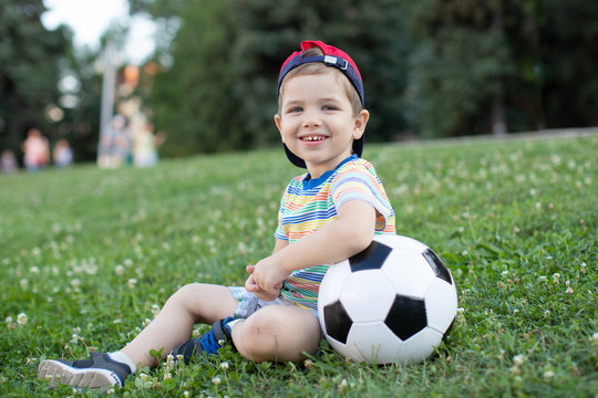 thoughtful little boy  sits on the grass and put his hands on a soccer ball