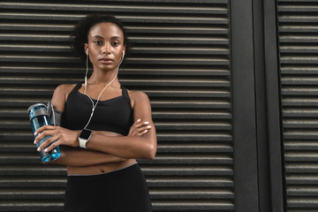 African american woman in fitness sportswear taking a break from excercises standing with a water...