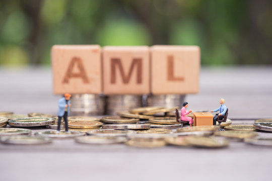 Money laundering concept. Miniature people, bank officer and customer on pile of coins and wooden word block "AML" as backdrop.