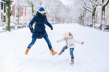 Fototapeta na wymiar Beautiful little girl wearing in jeans, white down jacket and knitted hat jumping with mama in snow outdoors. Having fun. Winter season. Childhood. Family winter vacation with child