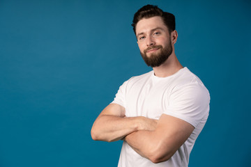 Fototapeta premium Charming handsome. Handsome young man in casual wear keeping arms crossed and smiling while standing isolated on blue background. Add space.