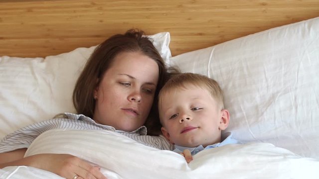 Close-up of sleepy mom and son lying on white pillows on the bed in the bedroom, they are covered with a warm blanket. Slow motion. Portrait of mother and son in bed in the morning.