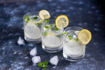 Pear tincture in a transparent glass. Ice cubes and pieces of fruit are also in the middle of the dishes. All on a dark background. Slices of lemon and mint leaves serve as a decoration for the drink.