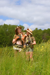 Two young girls seem to be on safari.They observe  the outdoor countryside area. They are dressed with  safari hats and khaki safari clothes.
