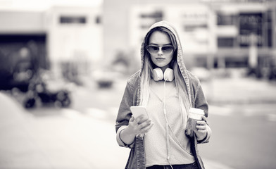 Young woman with headphones and sunglasses drinking coffee outdoor while texting on a smart phone. Urban style. Velvet Silver Retro