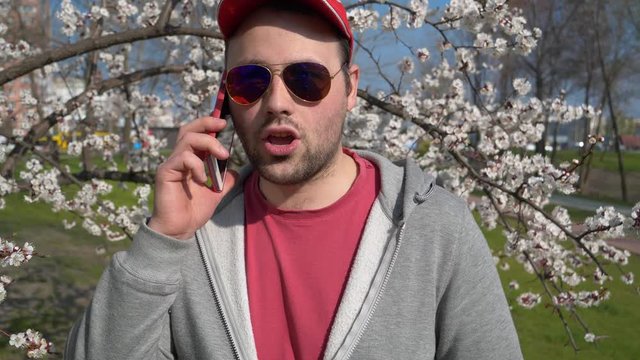 Man in sunglasses talks on smartphone in city park. Man in red baseball cap with red smartphone