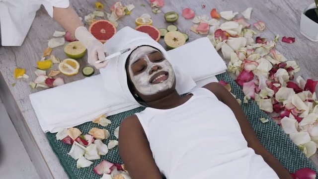 Top view of pretty young african woman relaxing while cosmetologist makes procedure of cleansing and moisturizing of skin in spa salon