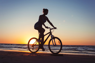 Naklejka na ściany i meble Silhouette of sporty woman riding bicycle on multicolored sunset background
