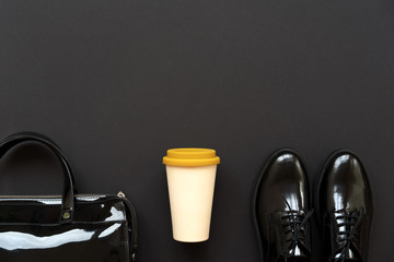 Black boots, a black lacquer bag and a bamboo cup with a yellow plastic lid for coffee to go on a black background. Concept back to school or back to work