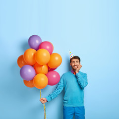 Fototapeta na wymiar Vertical image of nervous male bites finger nails, feels embarrassed in presence of many guests, poses with colorful balloons, celebrates birthday, wears cone paper hat. Student on leaving party