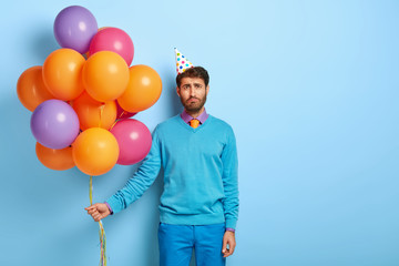 Studio shot of gloomy upset man looks unhappily at camera, feels lonely during his birthday, holds...
