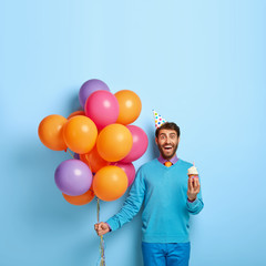 Happy birthday concept. Smiling European man holds little tasy cake, bunch of airballoons, going to...