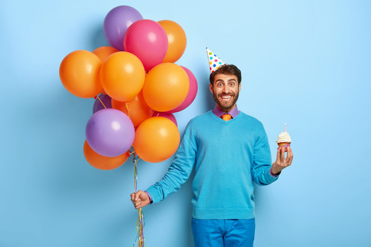 Glad young man with bristle, holds delicious small muffin, bunch of colored balloons, going to congraulate friend with birthday, dressed in blue clothes, party hat, models indoor. Holiday concept