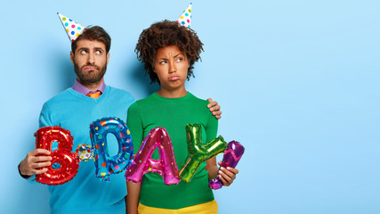 Photo of displeased mixed race couple embrace while hold colorful balloons meaning birthday, being in low spirit during celebration, make photo together, pose against blue studio wall. Copy space