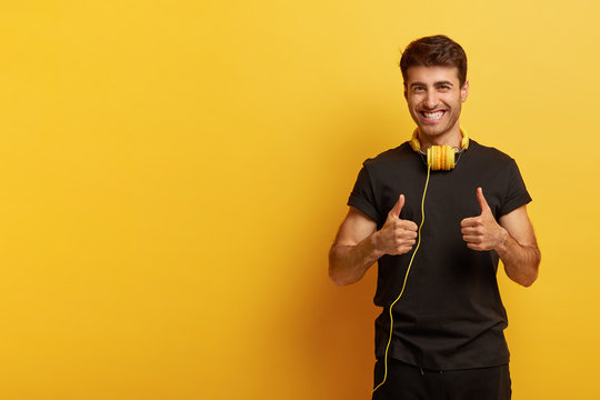 Yeah, I like this music! Positive hipster gives thumbs up, shows like gesture, expresses good emotions, wears black casual outfit, headphones on neck, isolated on yellow background, copy space