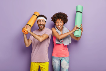 Horizontal shot of happy woman and man have fun after aerobics, raise hands with folded karemats, dressed in sportwear, enjoy spare time for sport, isolated on purple background. Diverse couple