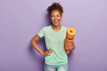 Overjoyed healthy dark skinned female athlete keeps hand on hip, holds rolled up fitness mat, being...