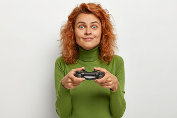 Cheerful adorable teenage girl focused with great interest, holds gamepad in hands, plays exciting game, wears green jumper, isolated on white background. Female plays video game on console station