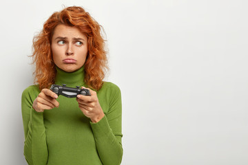 Sad hipster has red wavy hair, upset to loose game contest or challenge, holds playstation, being addicted gamer, dressed casually, has offended look, isolated on white background with copy space.