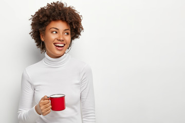 Joyful pleased dark skinned female drinks tea from red cup, looks on right side, glad to have spare...