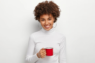 Portrait of happy curly haired woman with toothy smile, holds red mug of hot drink, looks straightly at camera, being coffee lover, dressed in white turtleneck jumper, has spare time, break after work