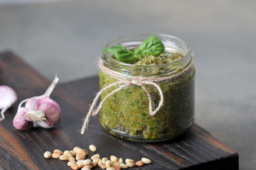 Glass jar with green pesto sauce. On a wooden board next to a jar of garlic, basil greens and pine nuts. Gray background. Close-up. Macro shot.
